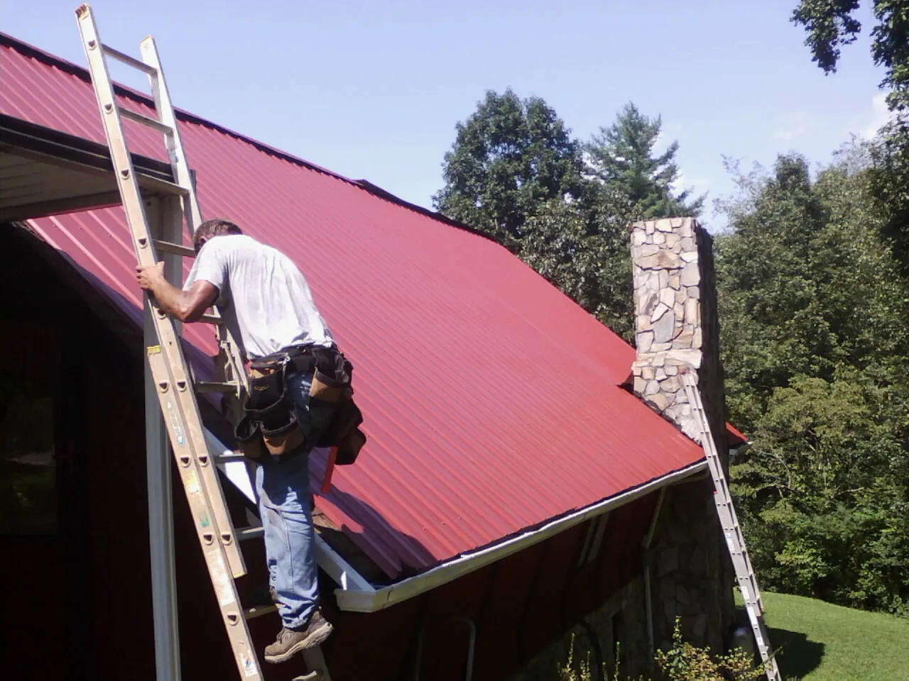 Roofer on a ladder beside a red metal roof and stone chimney — Soffit & Fascia Repair by Roofing Brothers in Bradner